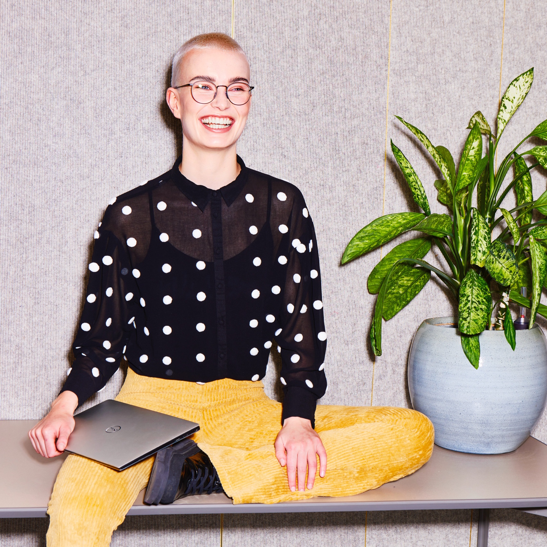 Een jonge, lachende vrouw met gemillimeterd witblond haar zit op een tafel naast een plant. Ze draagt een ronde bril, een zwarte blouse met witte stippen, en een gele ribfluwelen broek. In haar schoot houdt ze een dichtgeklapte zilveren laptop vast.